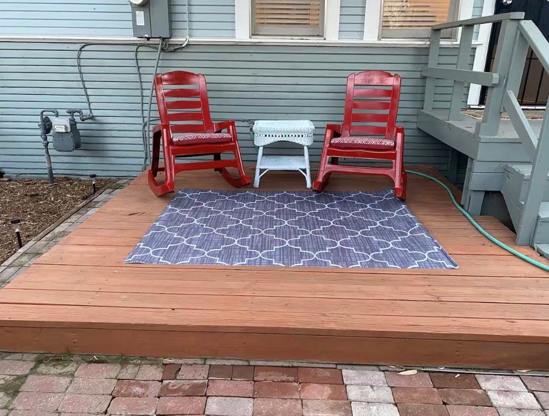 Una terraza de madera con dos sillas rojas y una mesita, un tapete estampado y un sendero de ladrillo que lleva a una casa.
