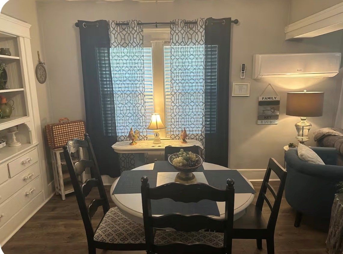 A dining area with a round table, chairs, a lamp, and a window with patterned curtains.