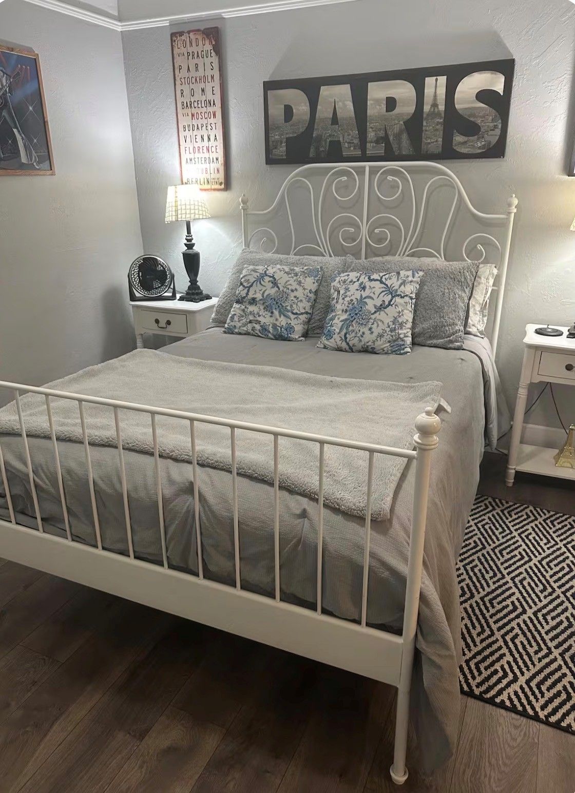 A bedroom with a white metal bed frame, a Paris-themed wall decoration, a bedside table with a lamp, and a patterned rug.
