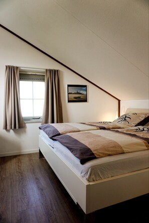 A bedroom with a bed, a window with curtains, a picture on the wall, and wooden flooring. - Your vacation home with a sauna and charging station on Texel – relaxation, nature, and the sea (De Cocksdorp)