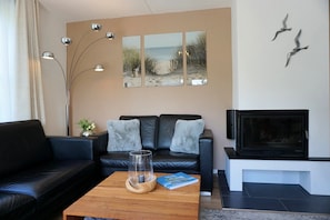 Ein Wohnzimmer mit einem schwarzen Ledersofa, einem Couchtisch aus Holz mit einem Buch, einem Kamin und Wandbildern, die eine Strandansicht zeigen.
