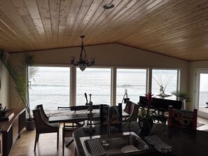 Une pièce spacieuse avec un plafond en bois, de grandes fenêtres donnant sur l’océan, un coin repas avec une table et des chaises, et une cuisine équipée d’un évier.