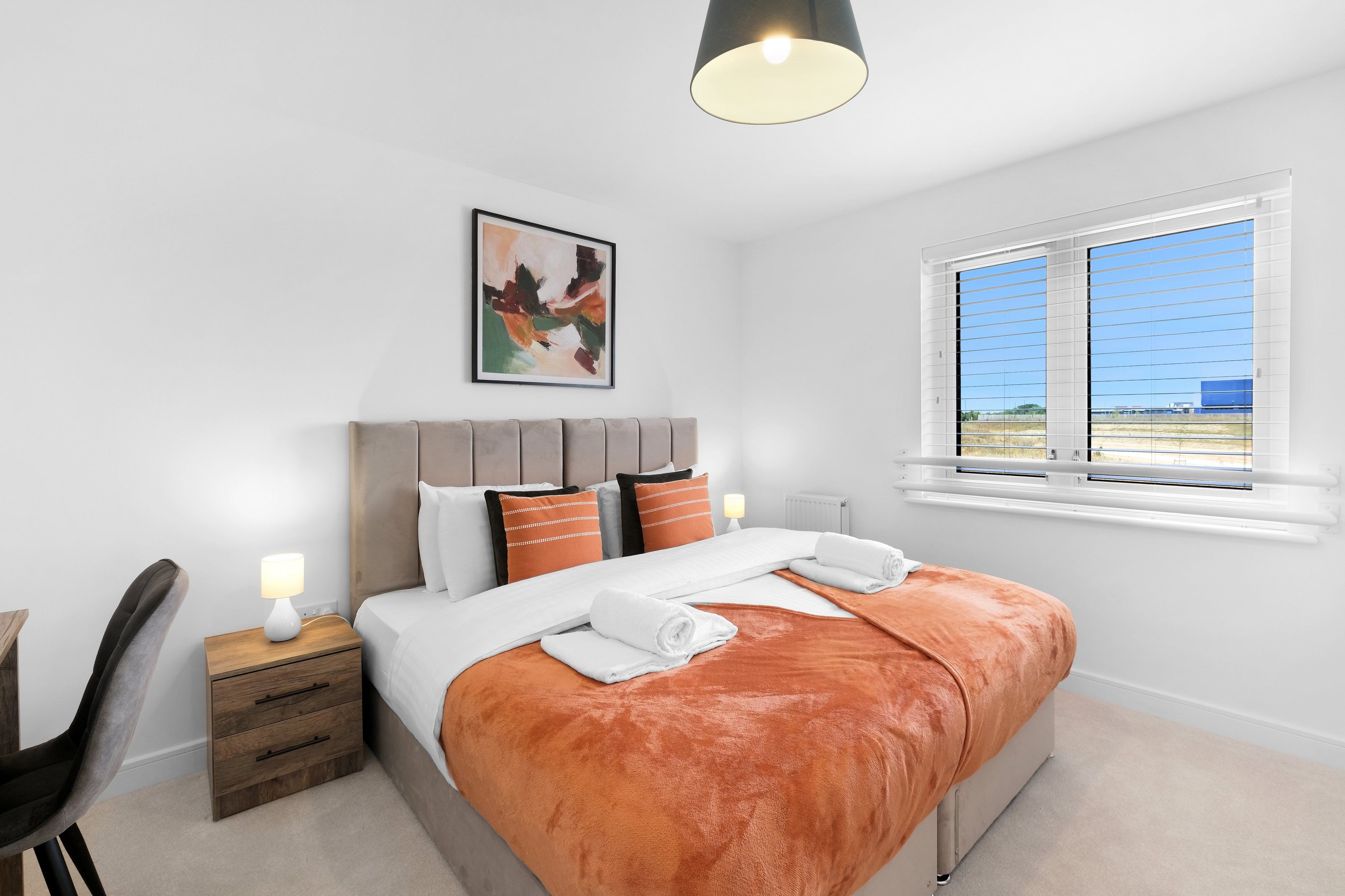 Quarto moderno com cama, mesas de cabeceira, uma cadeira, janela com persianas e luminária no teto.