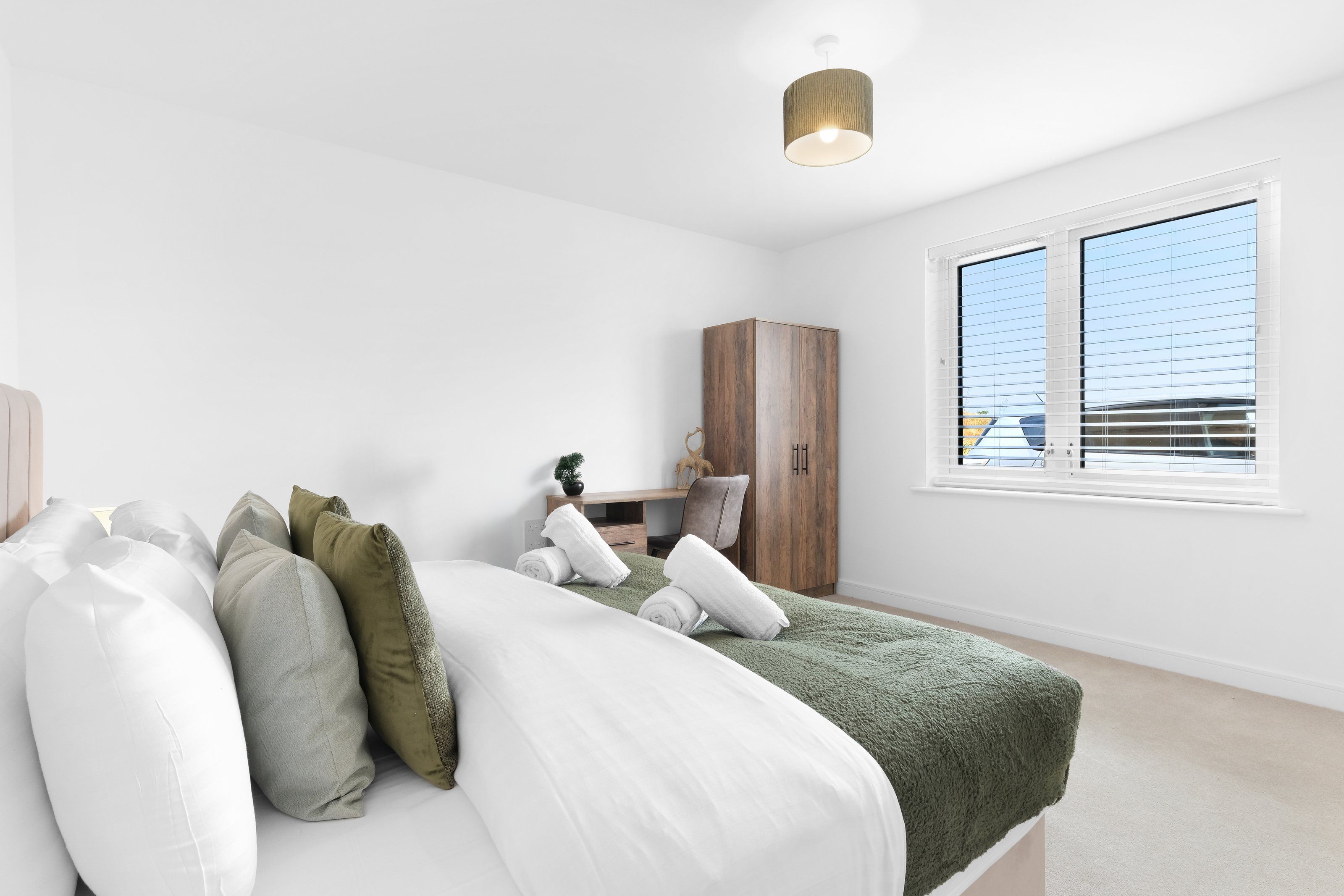 A bedroom with a bed, pillows, a nightstand, a wooden wardrobe, and a window with blinds.