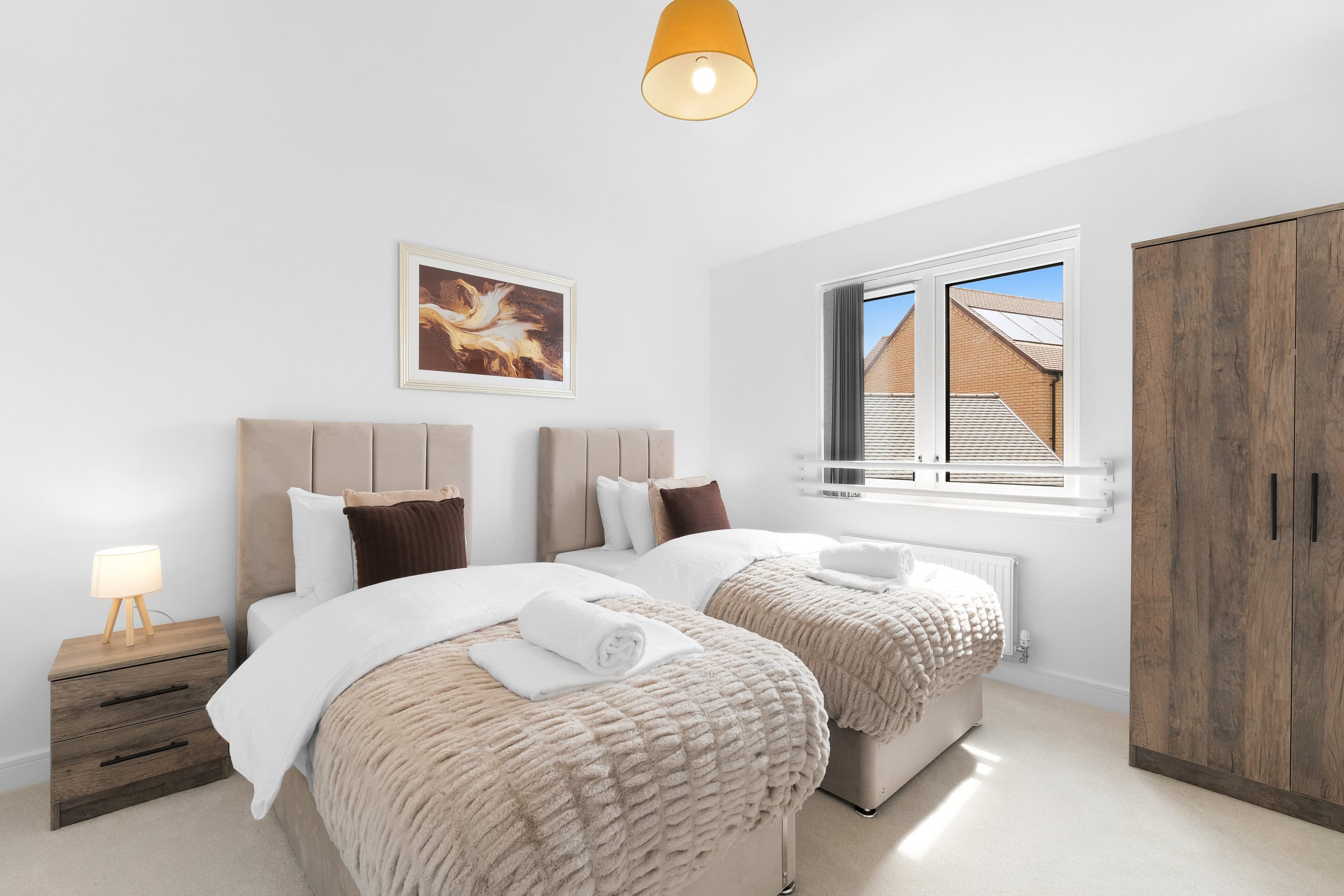 Een slaapkamer met twee bedden, een nachtkastje, een lamp, een lijst met een afbeelding, een raam met uitzicht en een garderobe.