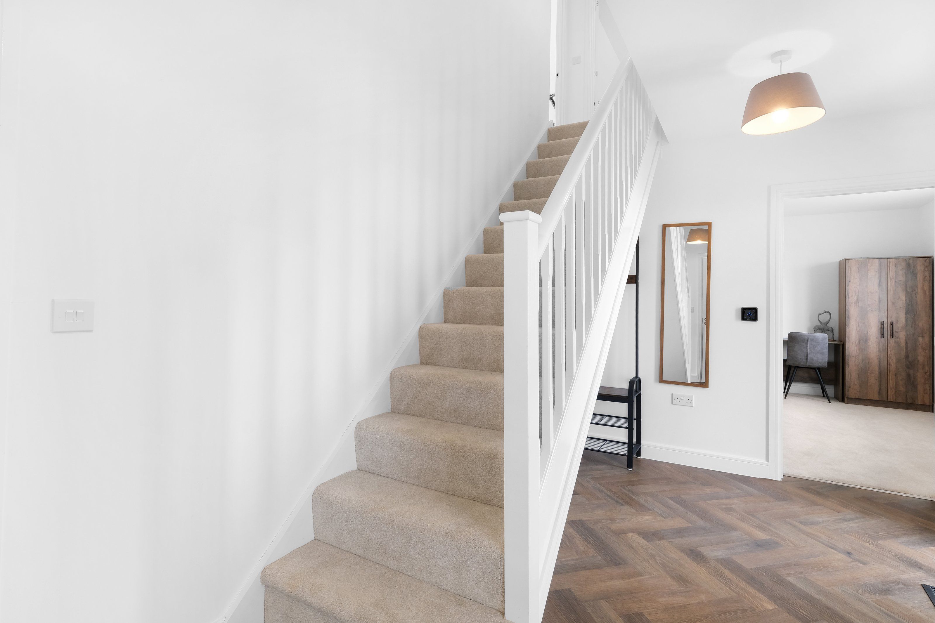 A staircase with beige carpeted steps and white railings, leading to a room with a herringbone wood floor and a wooden wardrobe.