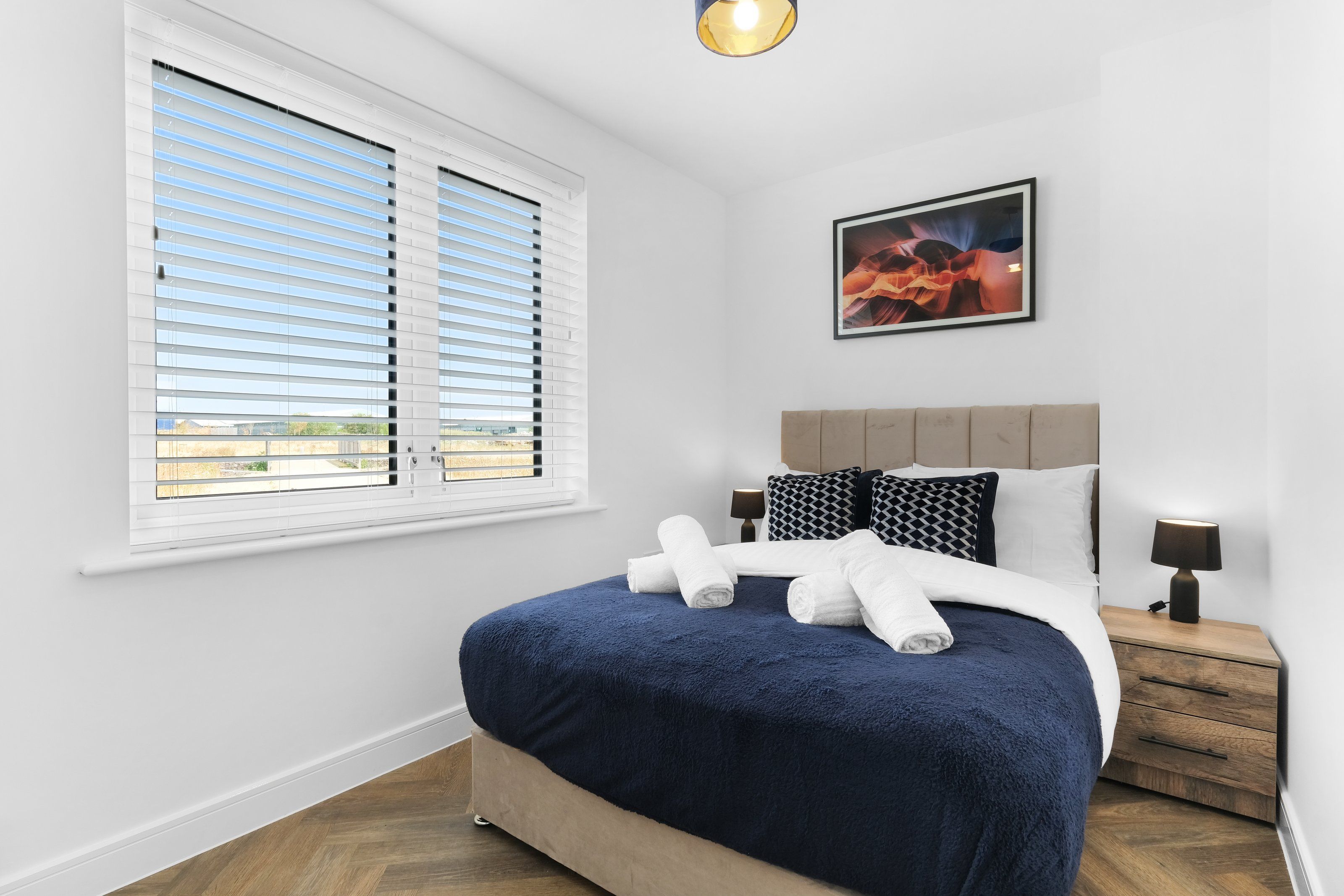A bedroom with a bed, a window with blinds, a nightstand with a lamp, and a framed picture on the wall.