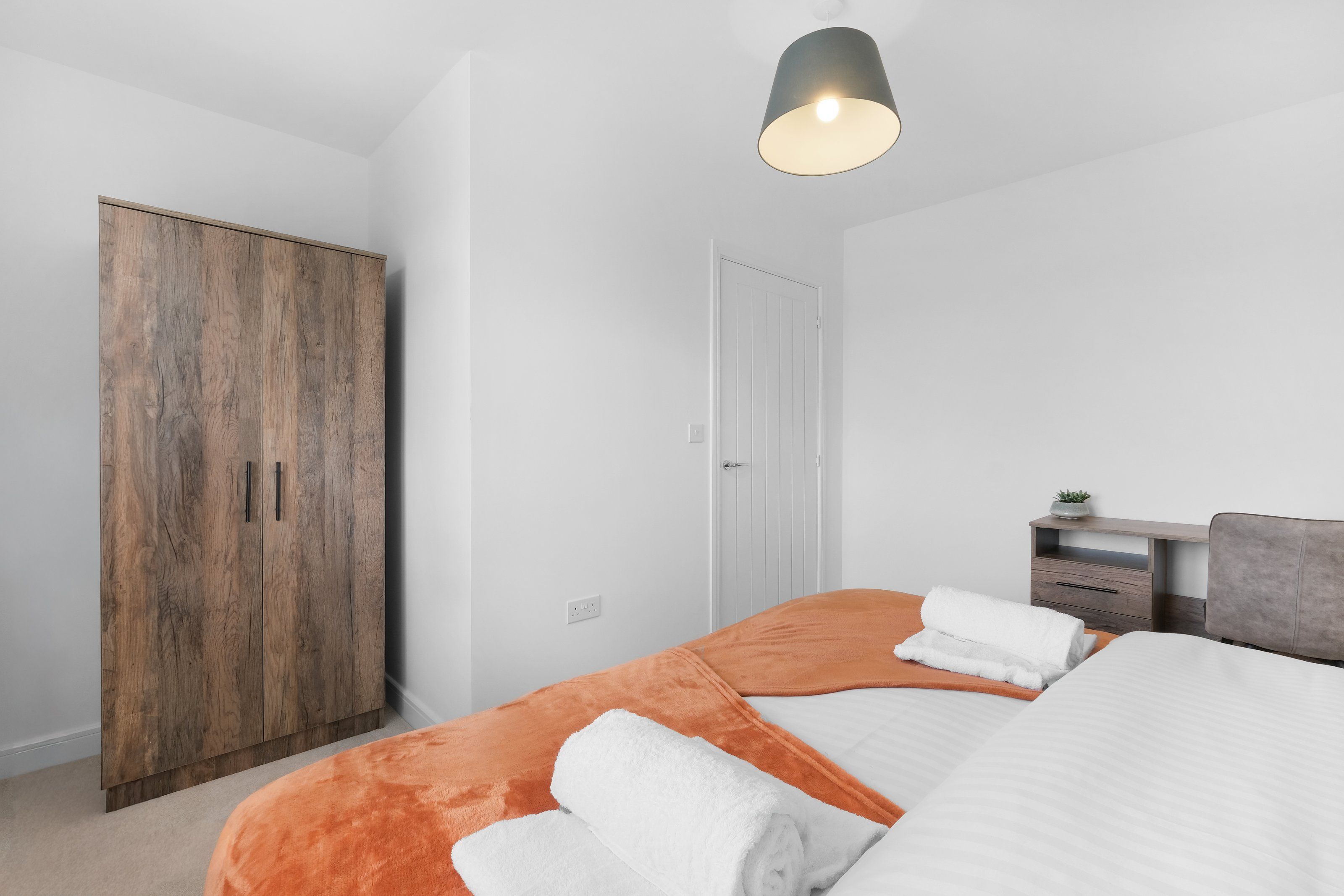 A bedroom with a bed, a wardrobe, a desk, and a hanging light fixture.
