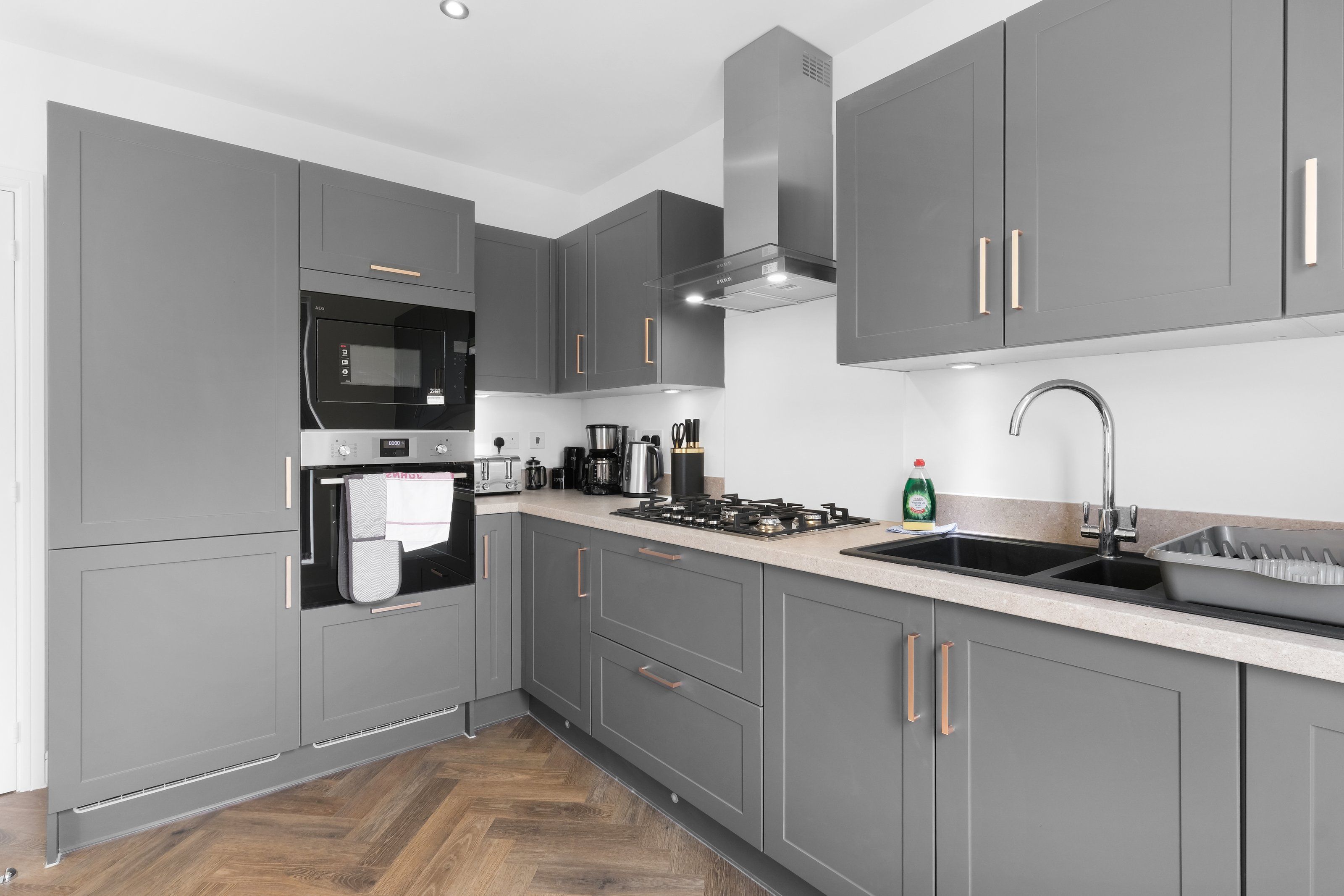 A modern kitchen with grey cabinets, stainless steel appliances, and a wooden herringbone floor.