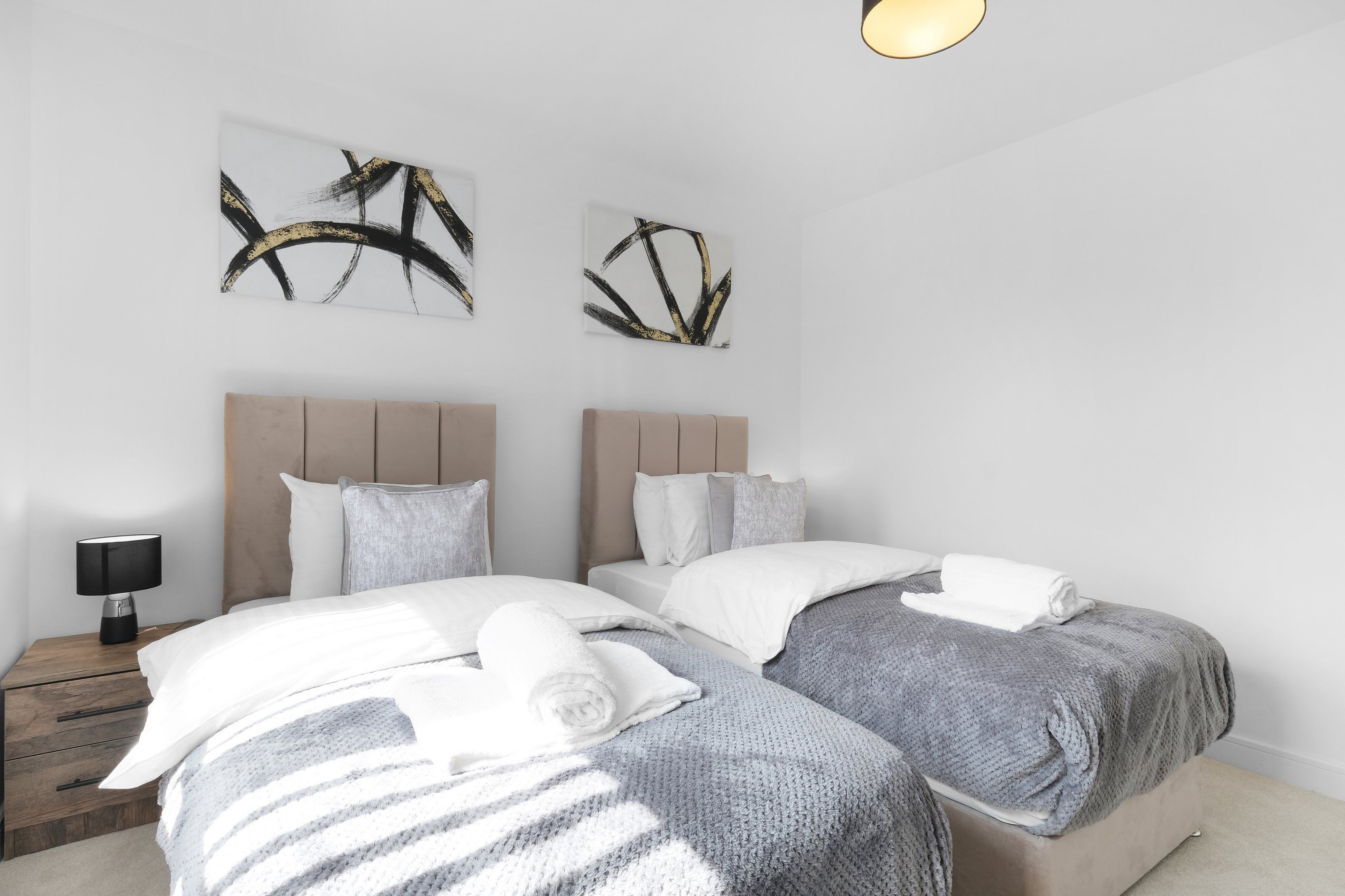 Hotellihuone, jossa on kaksi sänkyä, yöpöytä lampun kanssa ja abstraktia taidetta seinällä.