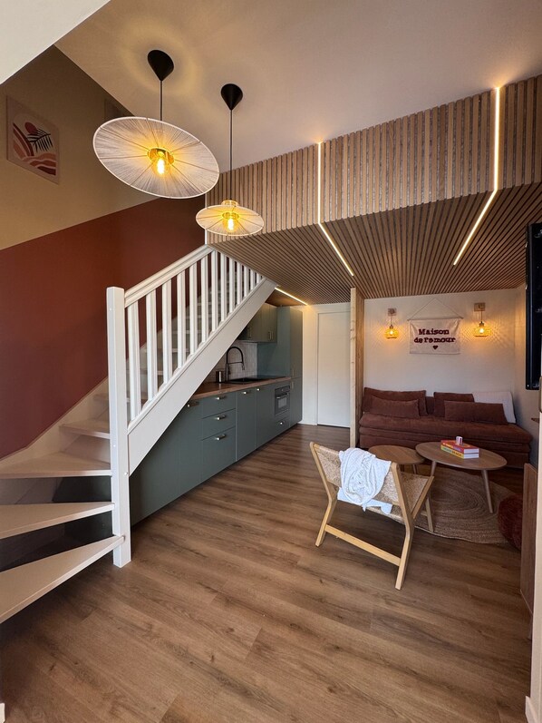 Un intérieur moderne avec un escalier, un plancher en bois, une cuisine, un coin salon avec un canapé et une petite table, ainsi que des luminaires suspendus.