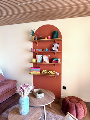 Un salon chaleureux avec un plafond en bois, une bibliothèque remplie de livres et d’objets décoratifs, une table basse ronde sur laquelle est posé un vase de fleurs, et un pouf rouge.