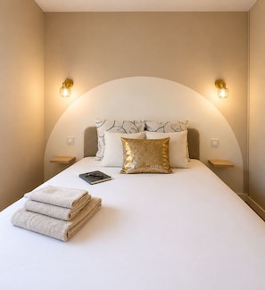 A neatly made bed with a white comforter, a gold pillow, and a pair of glasses on a book. There are two bedside tables with lamps, and the walls are painted in a neutral tone. - Monaco Luxury Apartment • Terrace • Parking • Sleeps 5. View of Monaco (Beausoleil)