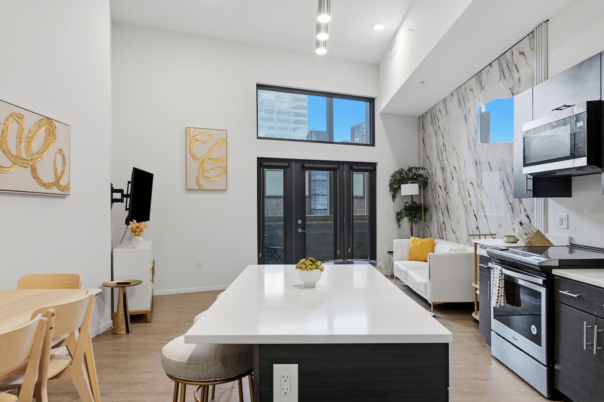 A modern kitchen with a central island, a dining area with a table and chairs, and a living space with a sofa and a plant.
