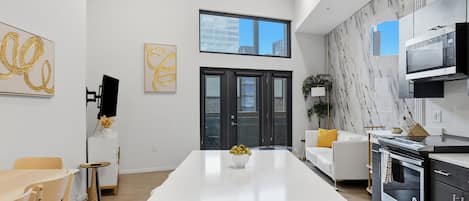A modern kitchen with a central island, a dining area with a table and chairs, and a living space with a sofa and a plant.