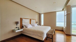 A bedroom with a large bed, bedside tables, a bench, and a view of the ocean.