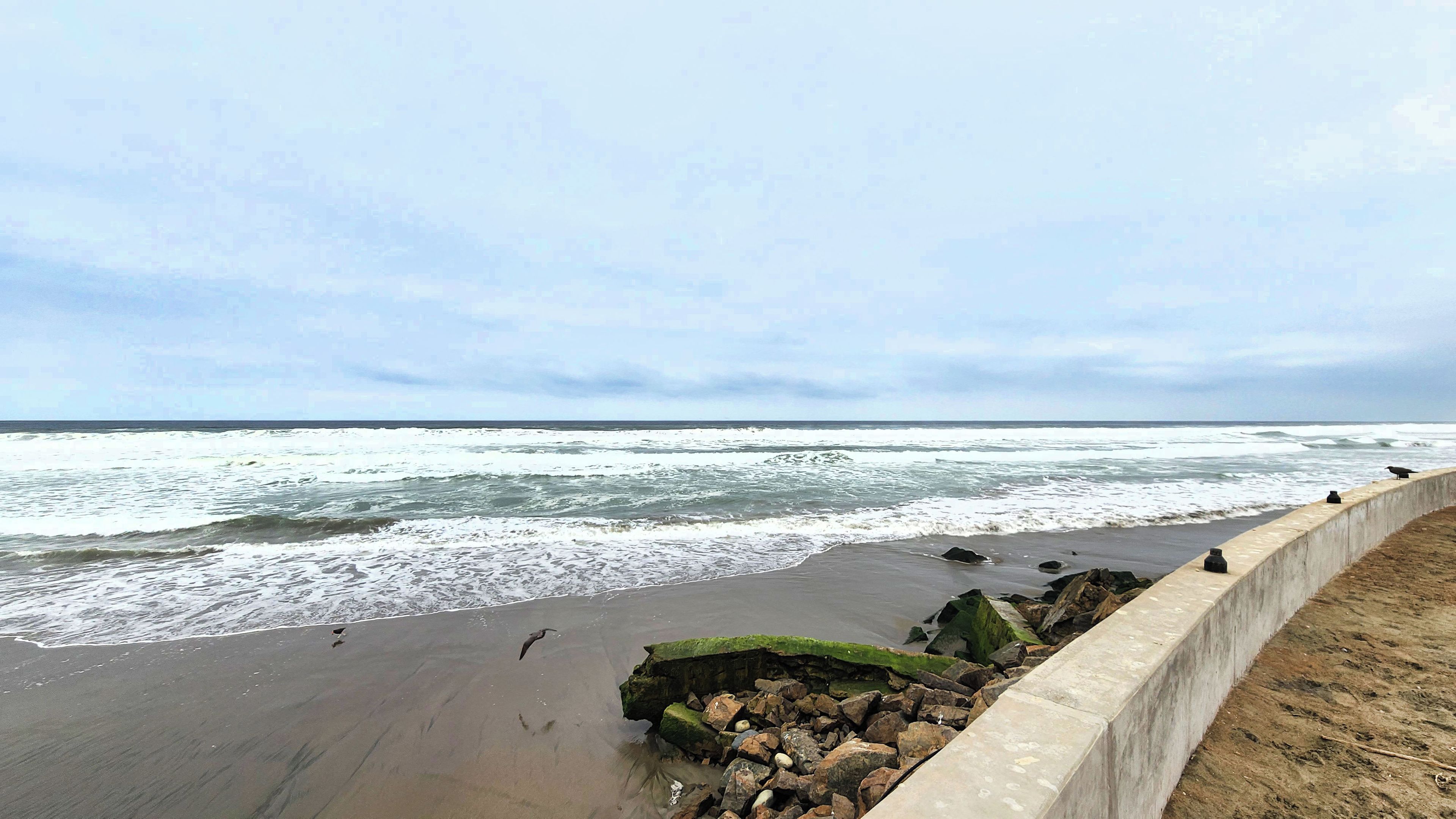Une plage avec des vagues, une barrière en béton et des rochers.
