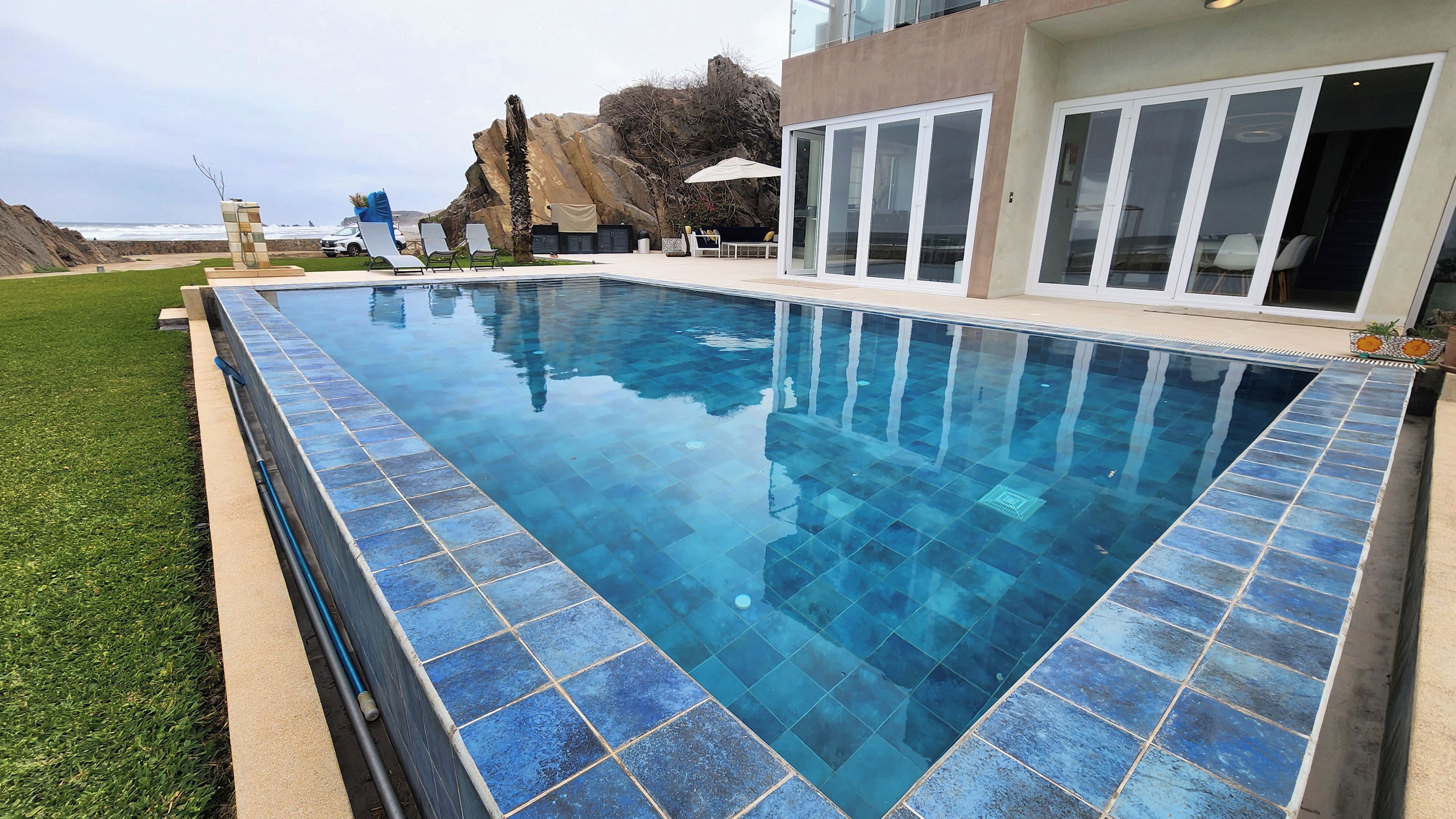 Une piscine rectangulaire aux carreaux bleus, entourée d’une pelouse et bordée d’un bâtiment moderne doté de grandes fenêtres.