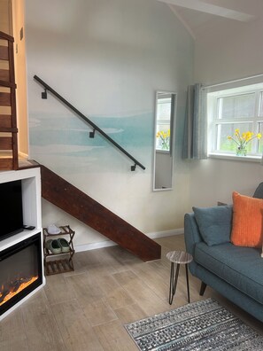 Een woonkamer met een open haard, een blauwe bank, een houten trap en een raam met uitzicht op het landschap.