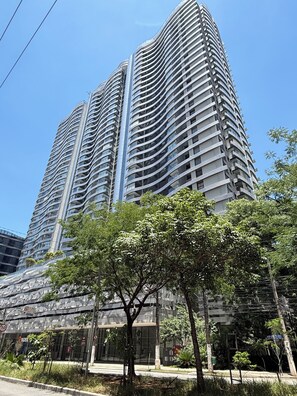 Um edifício alto com fachada curva, várias varandas e área comercial no térreo com uma árvore em primeiro plano.
