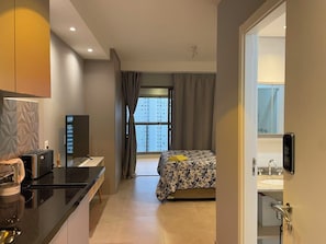 Quarto de hotel moderno com cozinha compacta, cama com colcha florida, grande janela com cortinas e banheiro com pia e espelho.
