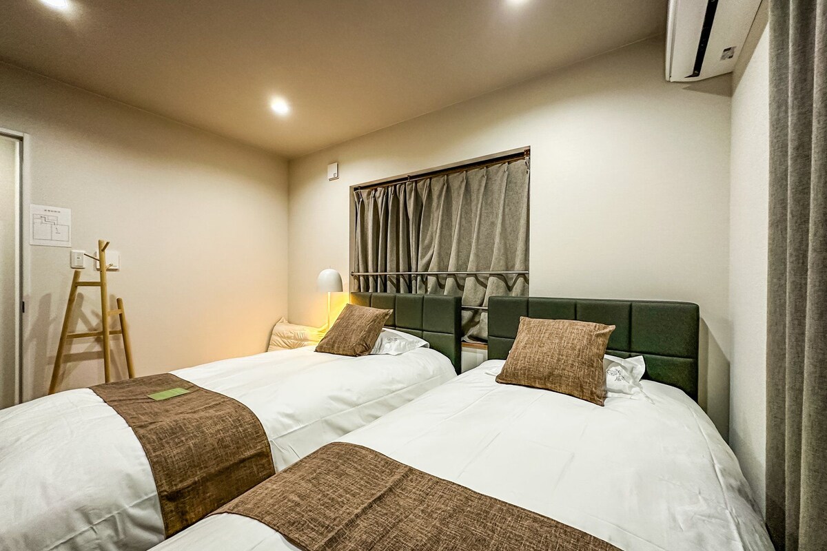 A hotel room with two beds, a window with curtains, a bedside lamp, and an air conditioning unit.