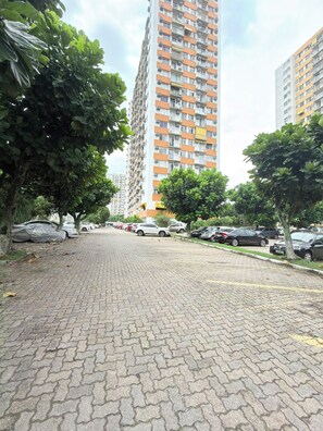 Un camino empedrado para peatones bordeado por coches estacionados y árboles, que conduce a un edificio residencial alto con varios balcones.