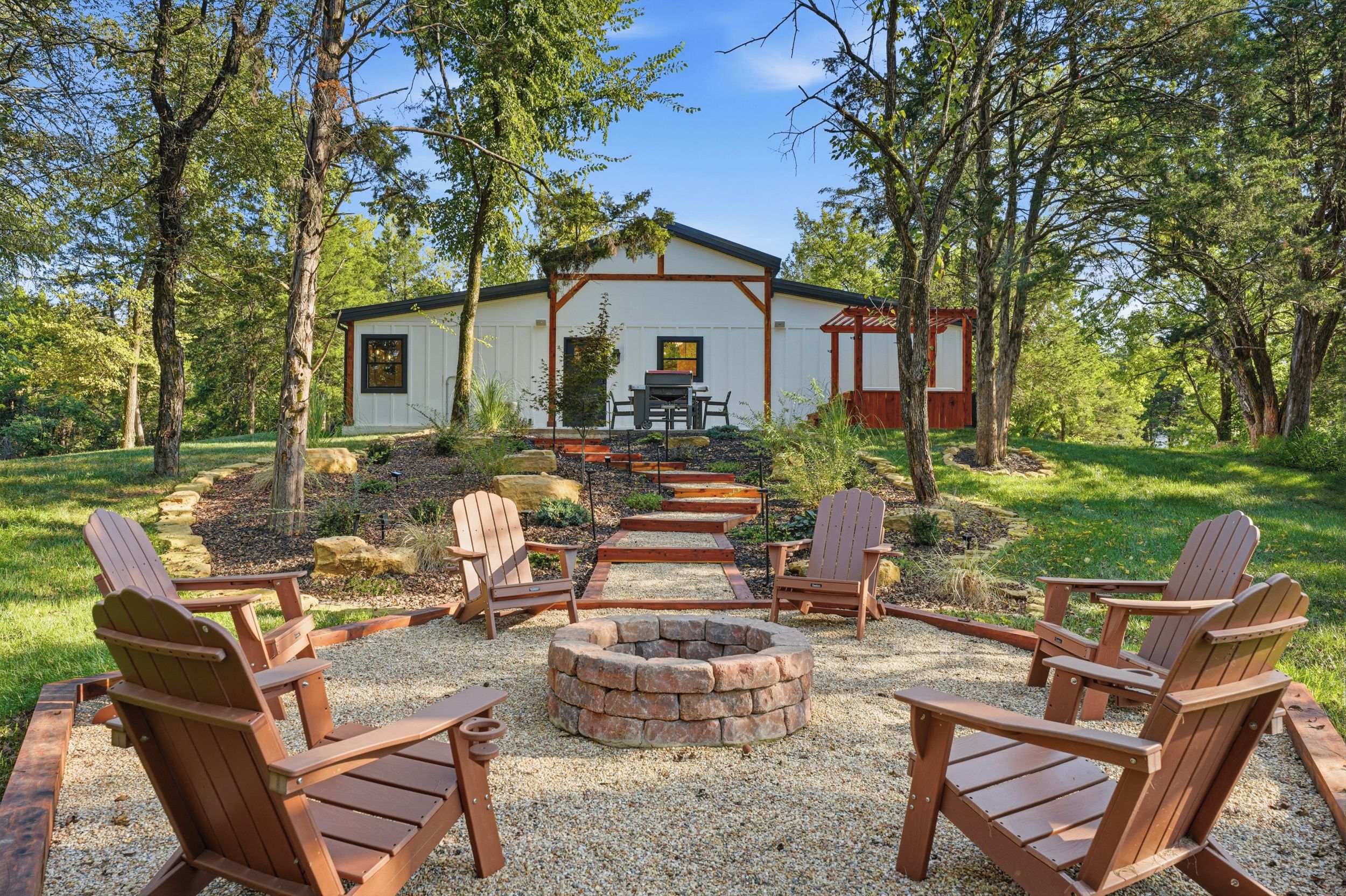 Une maison dotée d’une terrasse, entourée d’arbres, et un foyer avec des chaises disposées devant.