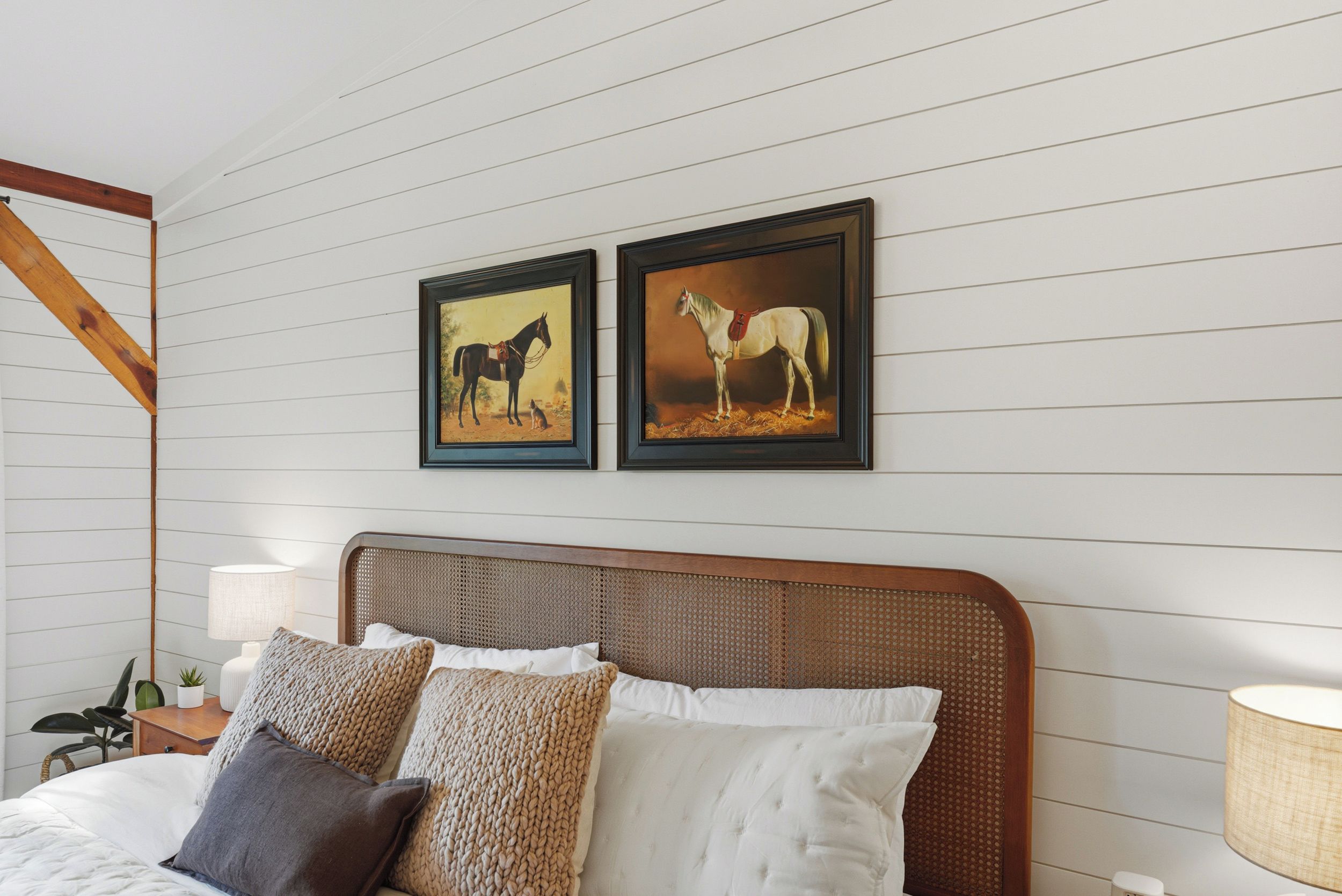 Quarto com cabeceira de madeira, cama com roupa de cama branca e várias almofadas, duas pinturas de cavalos emolduradas na parede e uma mesa de cabeceira com abajur.