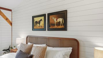 Quarto com cabeceira de madeira, cama com roupa de cama branca e várias almofadas, duas pinturas de cavalos emolduradas na parede e uma mesa de cabeceira com abajur.