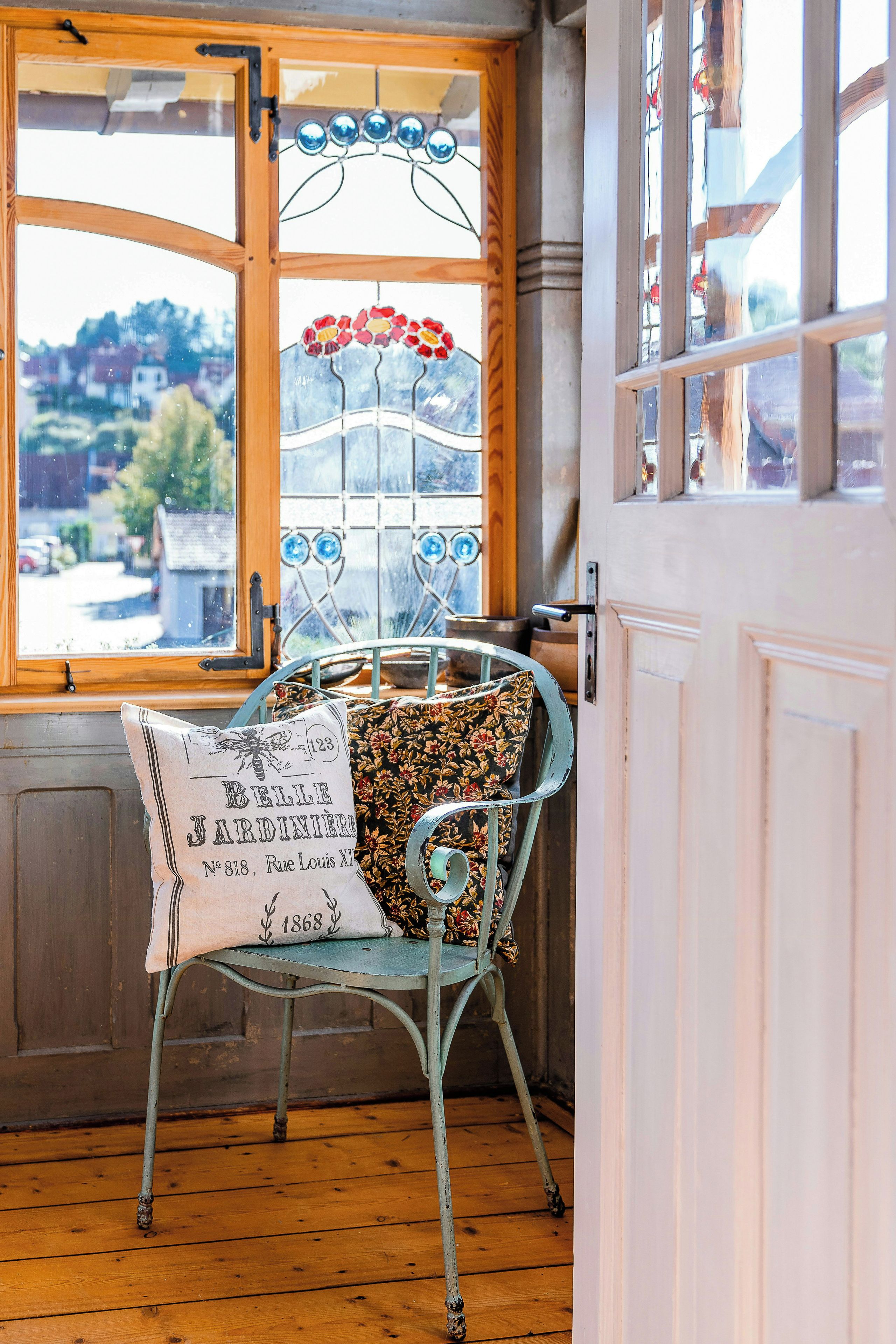 A vintage metal chair with a floral cushion in a room with a wooden floor and a window with a stained glass design.