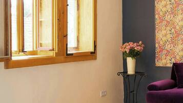 Una stanza con un letto, una finestra con persiane di legno, un tavolino con un vaso di fiori e una sedia viola.