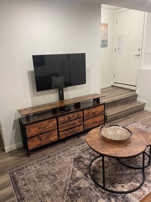 Una sala de estar moderna con un televisor de pantalla plana montado en una consola de madera, una mesa de centro redonda de madera con un tazón, y una alfombra estampada en el suelo.