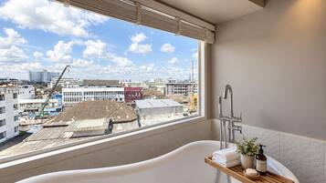 Ein modernes Badezimmer mit einer freistehenden Badewanne, einem Fenster mit Blick auf die Stadt und einer kleinen Holztrennwand für Toilettenartikel.