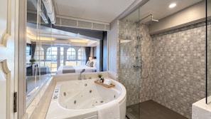 Un baño moderno con una bañera grande, ducha con mampara de vidrio y vistas a un dormitorio con cama y ventana.