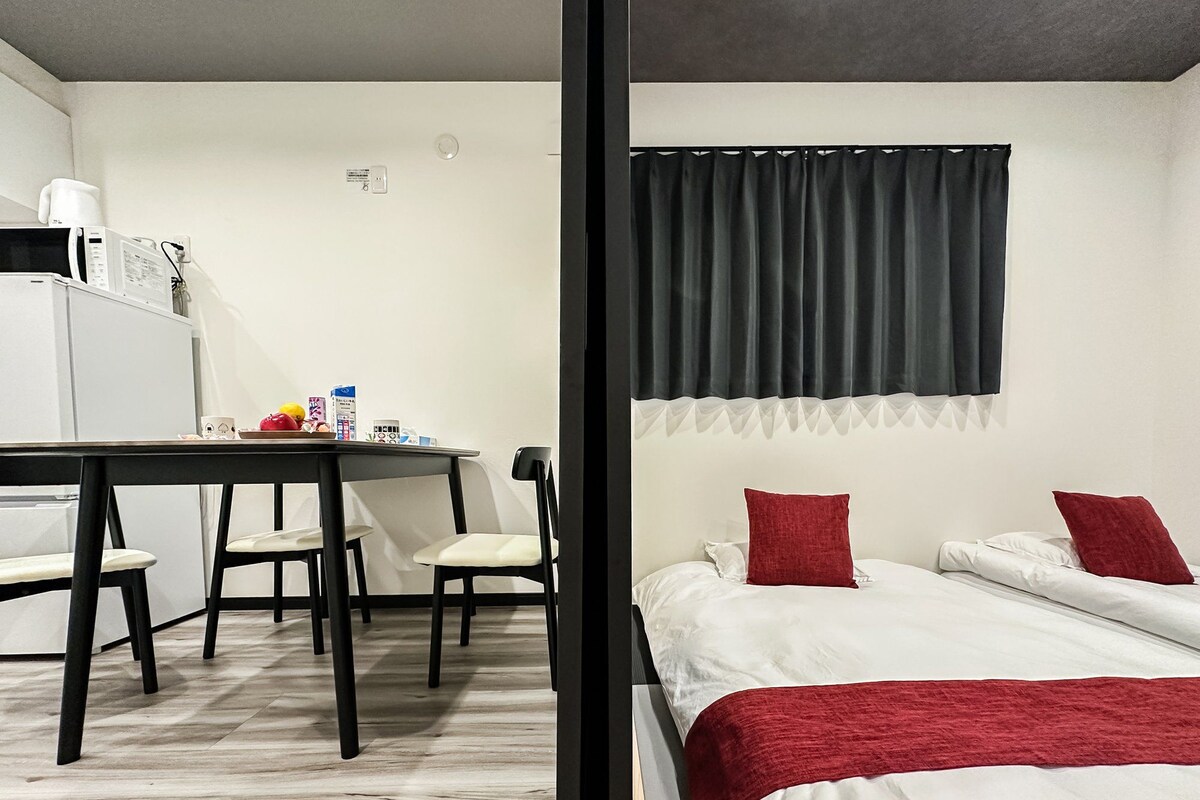 Ein kompaktes Hotelzimmer mit einer Küchenzeile, einem Bett mit roten Kissen und einem Fenster mit einem schwarzen Vorhang.