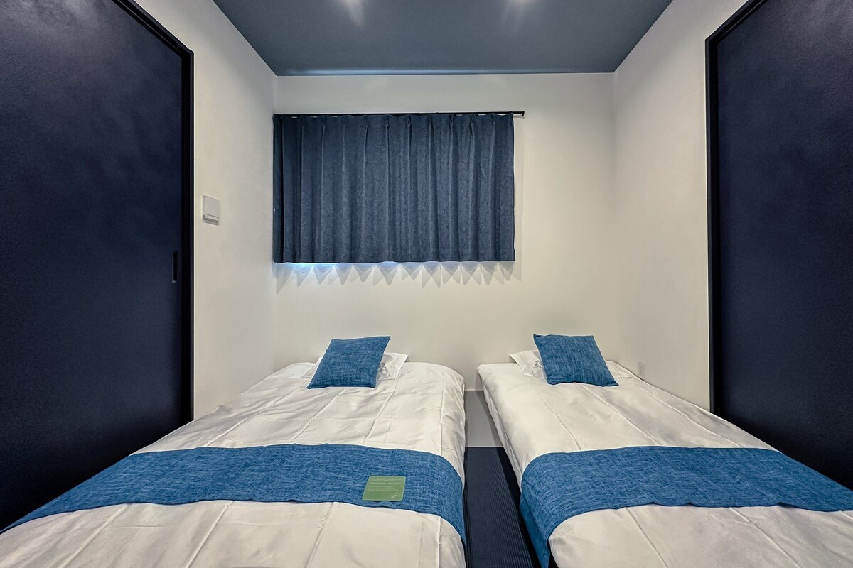 Ein Zimmer mit zwei Betten, jeweils mit blauer Decke und Kissen, einem Fenster mit blauer Vorhang und einer Deckenleuchte.