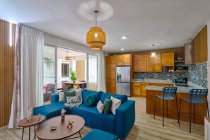 A modern living room with a blue sofa, a dining area, and a kitchen with wooden cabinets. - Luxury Gateway 2BR Bayahibe Pool Beach (Bayahíbe La Romana)