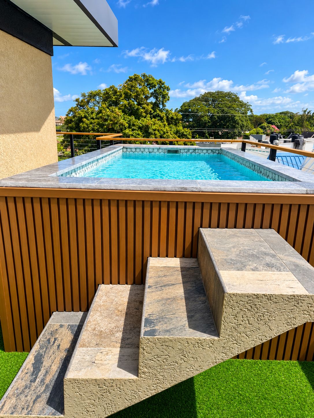 A swimming pool with a wooden deck and steps leading to it, surrounded by greenery.