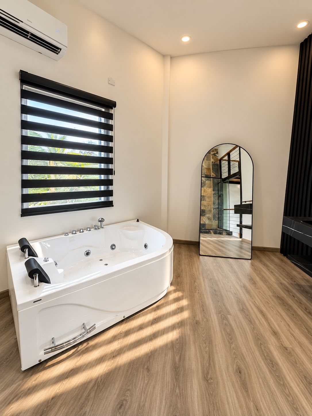 A modern bathroom with a freestanding bathtub, a window with blinds, a mirror, and a wooden floor.