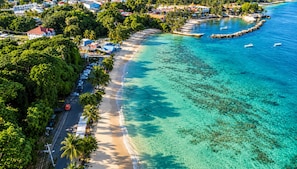 Une zone côtière avec une plage, des eaux cristallines et une route bordée de voitures garées et de palmiers.