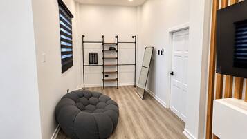 Un pasillo con suelo de madera, un sillón informal gris, una estantería metálica, un espejo apoyado contra la pared y una puerta.