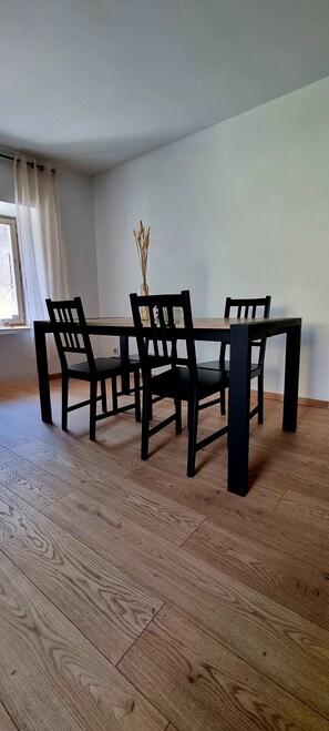 Ein Essbereich mit Tisch und vier Stühlen, einer Vase mit Trockenpflanzen und einem Fenster mit Vorhängen.