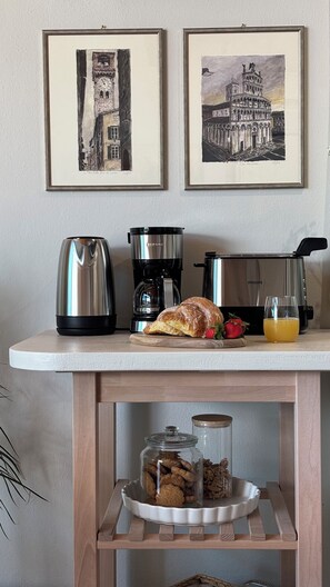 A kitchen counter with a coffee maker, a toaster, a croissant, strawberries, and a glass of orange juice. Below the counter, there are jars of cookies and a plant. - Almond Vista, Lucca Roof Garden (Lucca)