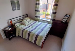 Een slaapkamer met een bed, nachtkastje, kapstok en een raam met gordijnen.