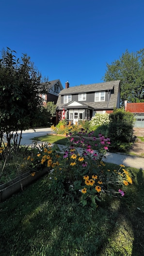 Ein zweistöckiges Haus mit Veranda, umgeben von einem Garten mit verschiedenen Blumen und einer Flagge.