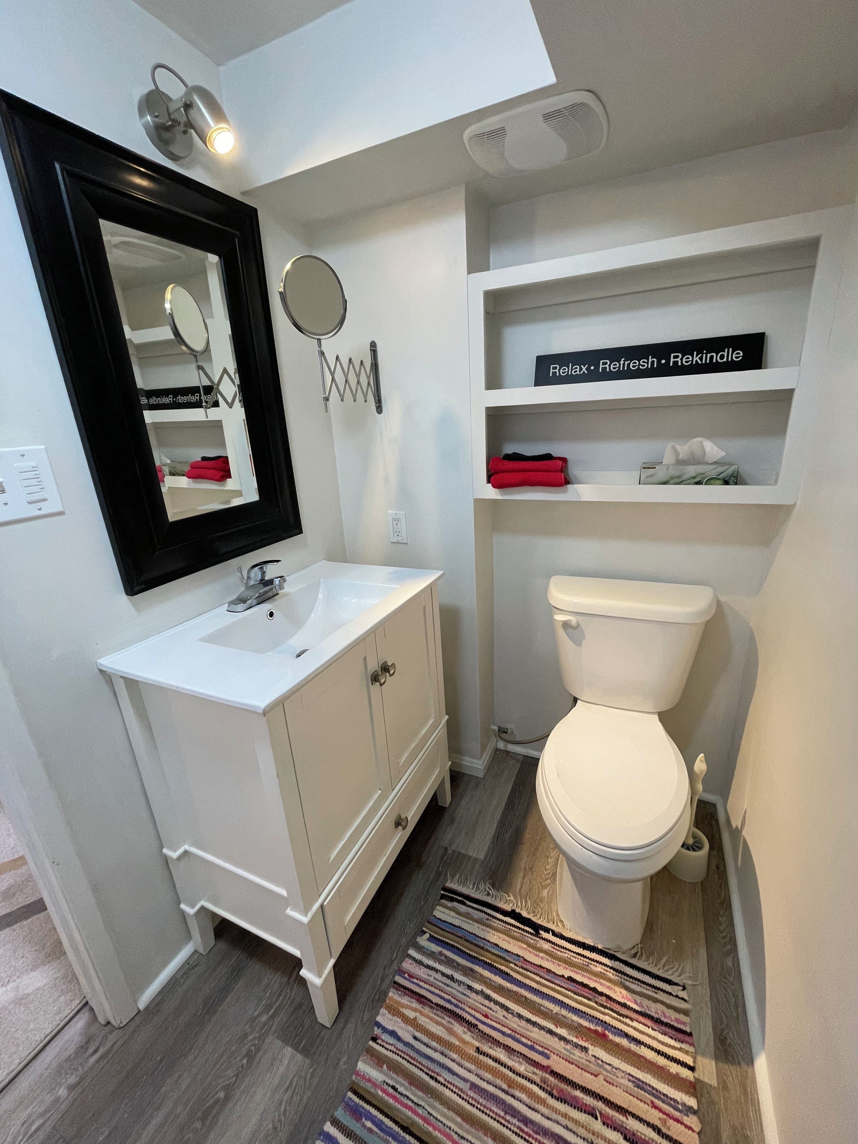 Une salle de bain avec un lavabo blanc, un miroir et des toilettes, un tapis à rayures et une étagère sur laquelle est posé un panneau.
