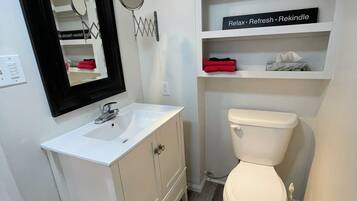 Une salle de bain avec un lavabo blanc, un miroir et des toilettes, un tapis à rayures et une étagère sur laquelle est posé un panneau.