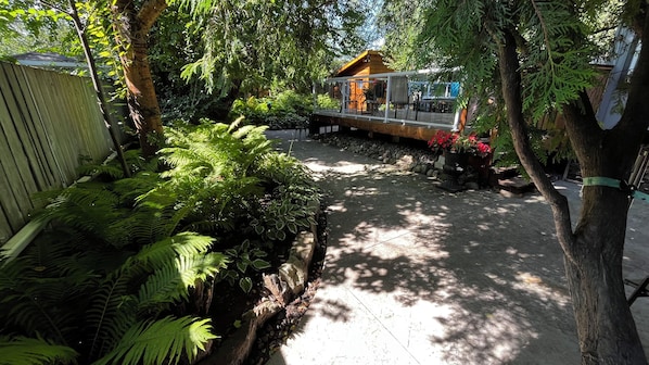 Un jardín con terraza de madera, plantas en macetas y un edificio con cerramiento de vidrio.