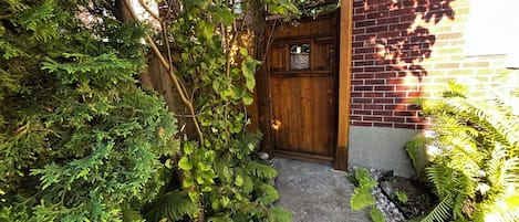 A wooden door with a small window, surrounded by dense greenery and a stone pathway leading to it.